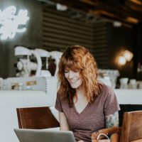 Red-haired woman using a laptop in a coffee shop, symbolizing remote DevOps work.
